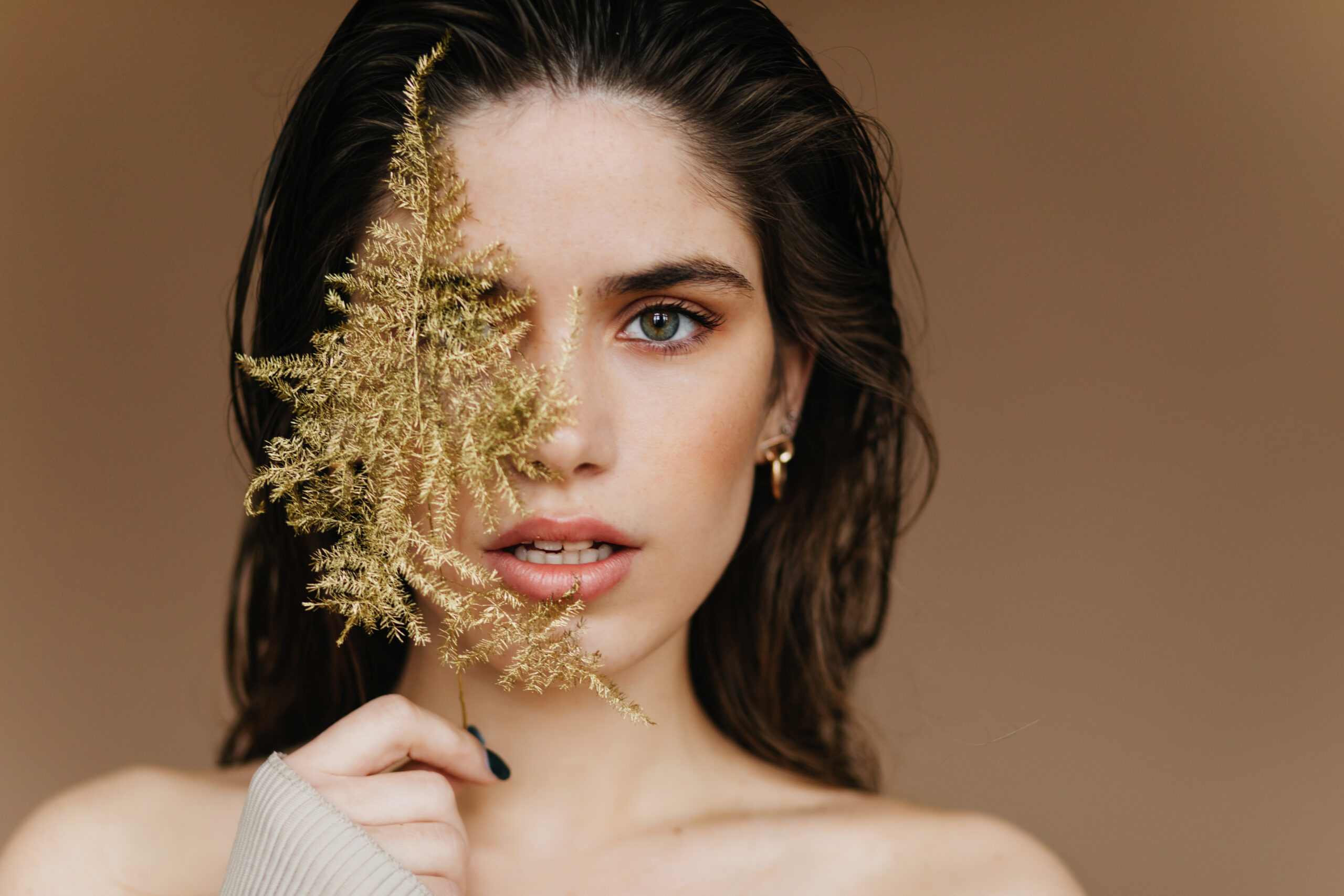 Elegant pale girl with golden earrings expressing amazement. Close-up shot of sensual brunette lady with plant.