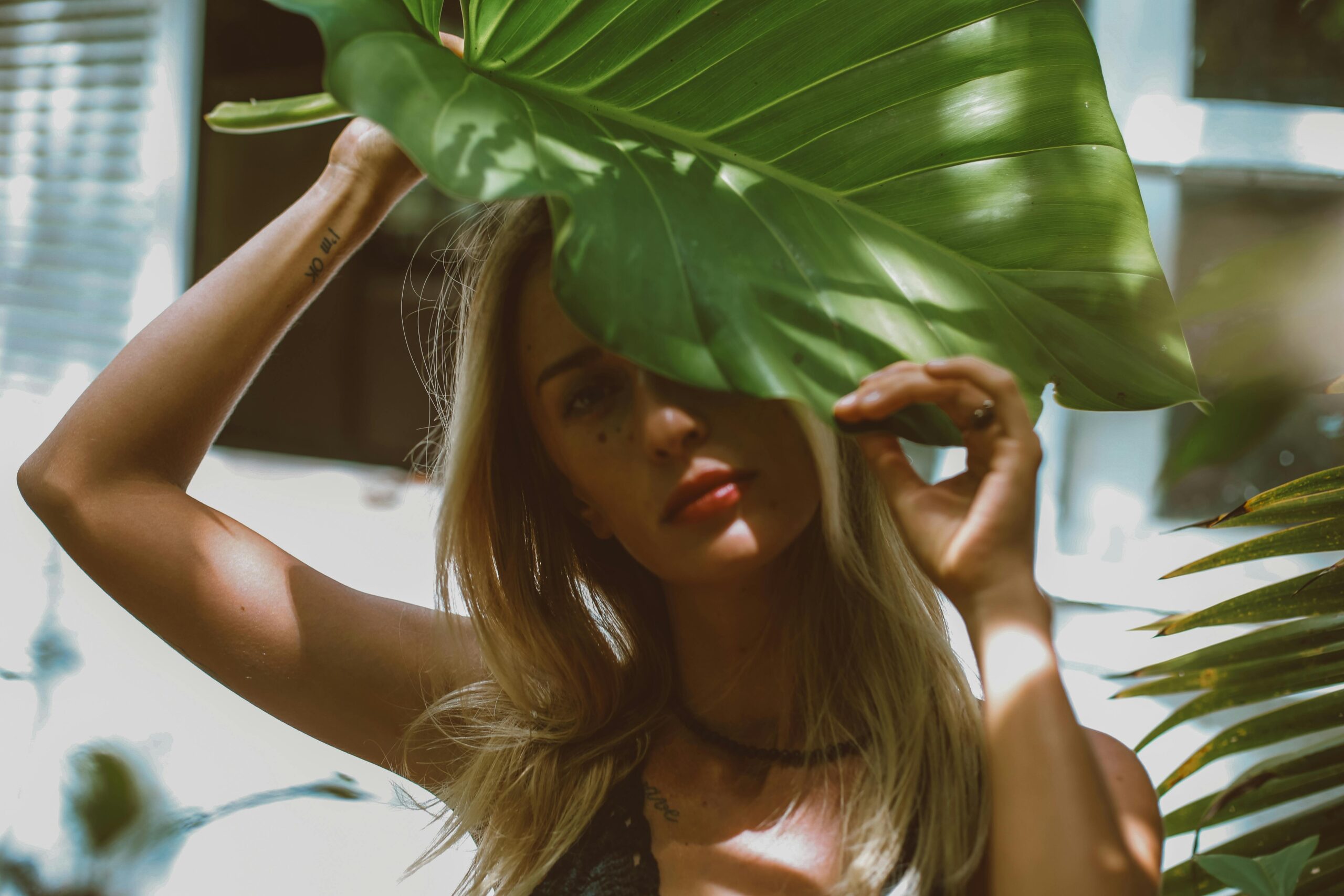 A serene portrait of a blonde woman partially covered by a tropical leaf, creating dramatic shadows outdoors.